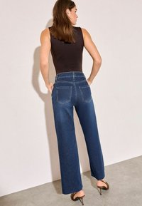 Jeans anchos de color azul oscuro con cintura alta y dos bolsillos traseros, combinados con una blusa ceñida negra y zapatos de tacón alto marrones.