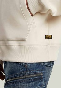 Beige sweatshirt met geribde zoom en G-Star Originals logo patch boven de blauwe spijkerbroek met een horizontale ritszak.