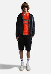 Zwarte lichtgewicht jack over een oranje hoodie, met een gestreept ontwerp en de Noorse vlag. Gecombineerd met zwarte cargoshorts en witte sneakers.