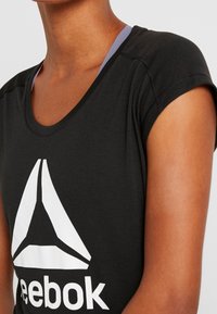 Černý sportovní top s krátkým rukávem, kulatým výstřihem a bílým logem Reebok. Má hladkou látku a kontrastní fialový pásek pod ním.