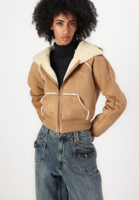Veste en daim marron avec un col en peau de mouton blanc, fermeture éclair sur le devant et deux poches avant. Associée à un jean en denim bleu taille haute.