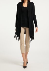 Cardigan noir ouvert à l'avant avec un ourlet à franges, porté sur un haut noir côtelé et associé à un pantalon beige raccourci et des escarpins noirs.