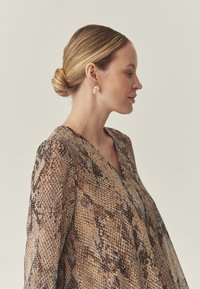 Braune Bluse mit Schlangenmuster, V-Ausschnitt, fließendem Stoff und langen Ärmeln, kombiniert mit runden Ohrringen in Goldoptik. Glatte Texturen und Muster sind sichtbar.