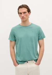 Light green cotton t-shirt with short sleeves, crew neckline, and subtle logo on the chest. Paired with light-colored pants.