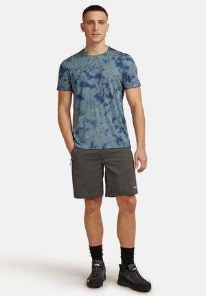 Hombre de pie con una camiseta azul tie-dye, pantalones cortos gris oscuro, calcetines negros y zapatos de senderismo negros sobre un fondo blanco liso.