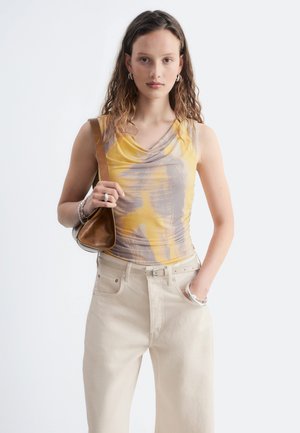 Jeune femme aux cheveux ondulés portant un débardeur jaune et gris, pantalon beige, tenant un sac à main marron avec la main dans la poche.