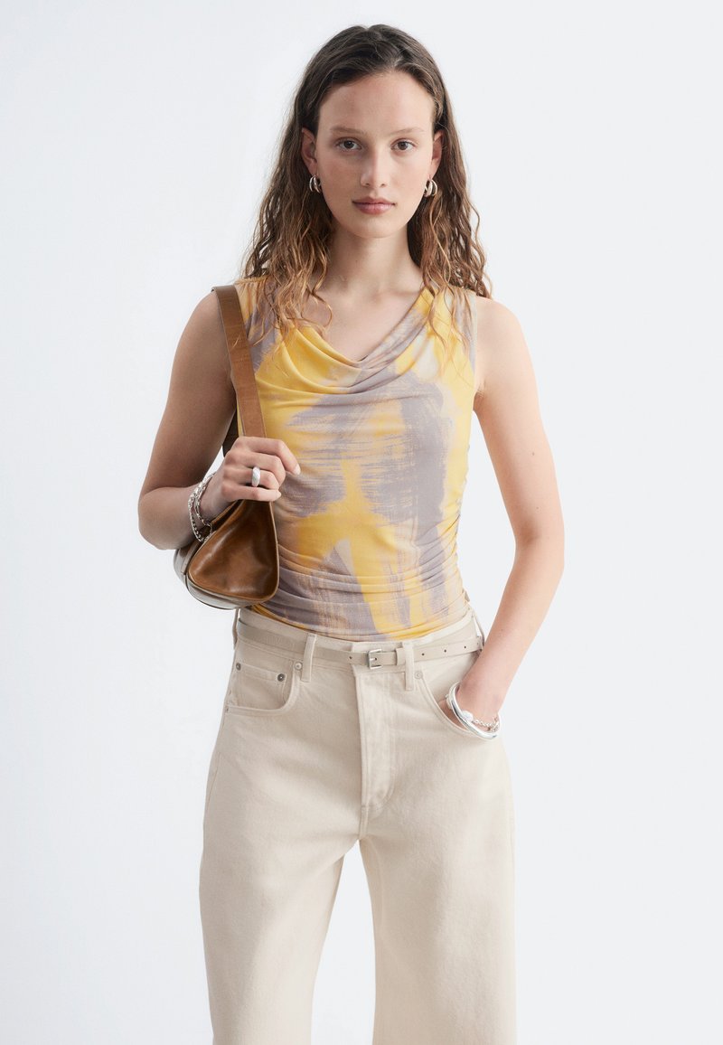 Jeune femme aux cheveux ondulés portant un débardeur jaune et gris, pantalon beige, tenant un sac à main marron avec la main dans la poche.