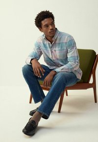 Camicia a quadri in azzurro chiaro, rosa e bianco; jeans in denim blu; mocassini neri; seduto su una sedia di metà secolo.