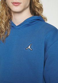 Osoba s kudrnatými blond vlasy má na sobě modrou mikinu s kapucí, na jejímž hrudníku je malé černobílé logo basketbalového hráče.