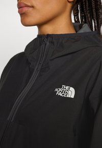 Közelkép egy személy arcáról, aki fekete kapucnis dzsekit visel, amelyen bal oldalon fehér "The North Face" logó található.