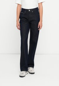 Tamsiai mėlyni džinsai su aukštu liemeniu, plačiais kirpimais ir priekinėmis kišenėmis. Apima metalinę mygtuko uždarymo sistemą ir subtilius siuvinėjimo akcentus.