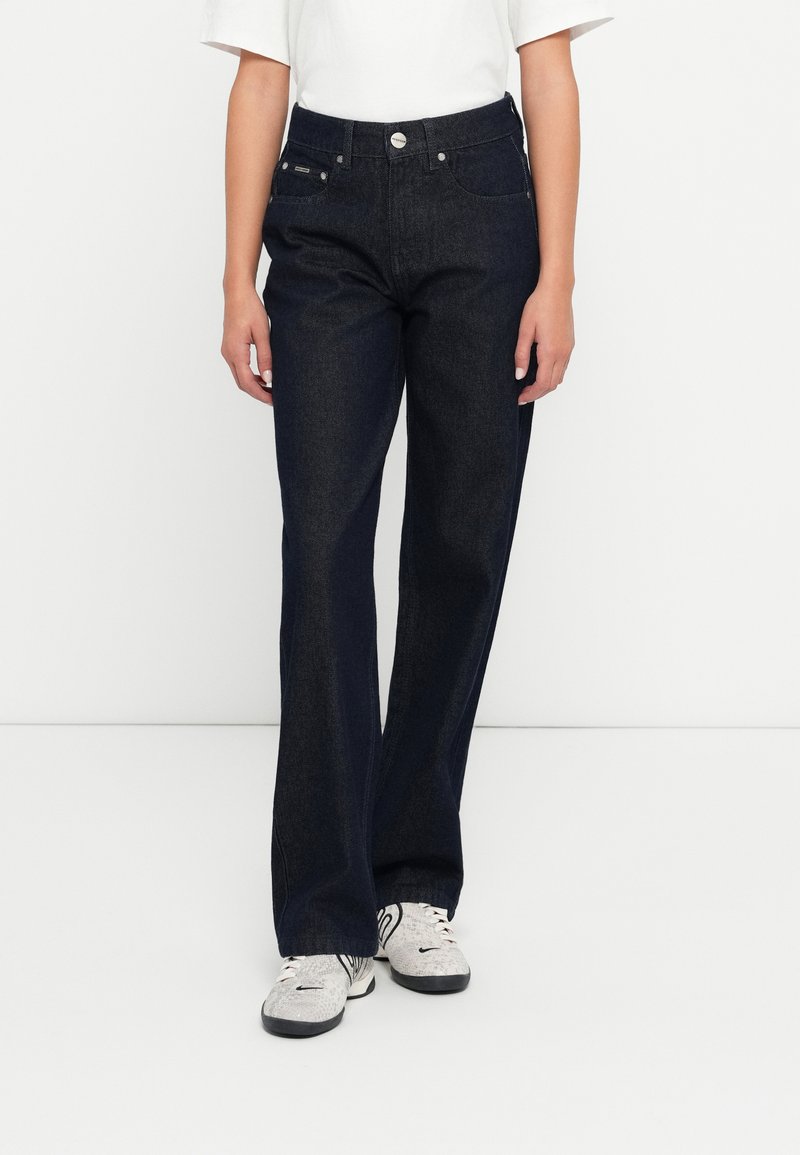 Tamsiai mėlyni džinsai su aukštu liemeniu, plačiais kirpimais ir priekinėmis kišenėmis. Apima metalinę mygtuko uždarymo sistemą ir subtilius siuvinėjimo akcentus.