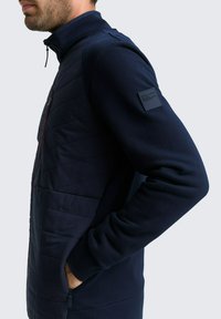 Marineblaues Jackett mit gesteppten Vorderpanelen, hohem Kragen und einem kleinen Logopatch am linken Ärmel. Mit Reißverschluss versehenes Taschen-Detail.