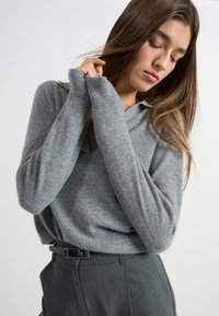 Szary sweter dzianinowy z dekoltem w kształcie V i długimi rękawami, z prążkowanymi mankietami. W zestawie z ciemnoszarymi spodniami od garnituru i czarnym paskiem.