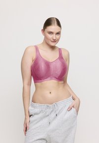 Nainen, jolla on vaalea iho ja vaaleat hiukset poninhännällä, pukeutuu vaaleanpunaiseen kuviolliseen rintaliiviin sekä vaaleanharmaisiin collegehousuihin, joista toisessa käsi on taskussa.