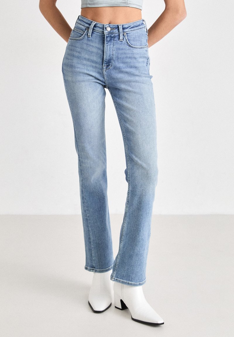 Jeans azzurri chiari a vita alta con gamba dritta, dotati di cinque tasche, leggera scoloritura e chiusura frontale con bottone. Indossati con stivali bianchi alla caviglia.