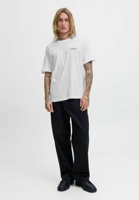 Jaunas vyras su ilga šviesia plaukus ir tatuiruotėmis, vilkintis baltą „Jack & Jones“ marškinėlį, laisvas juodas kelnes ir juodus batus, stovi prieš baltą foną.