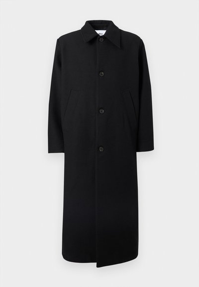 Μακρύ μαύρο παλτό με μυτερό γιακά, τέσσερα κουμπιά μπροστά και δύο κεκλιμένες πλαϊνές τσέπες σε λευκό φόντο.