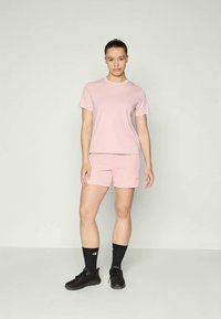 Moteris stovi, vilkėdama šviesiai rožinį trumpomis rankovėmis marškinėlį ir atitinkamas šortus, juodus sportinius batus ir juodas trumpas kojines.