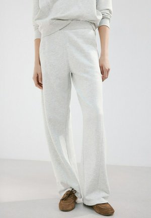 Persona vestida con pantalones deportivos sueltos de color gris claro, una sudadera a juego y zapatos marrones de ante con cordones, de pie frente a un fondo sencillo.