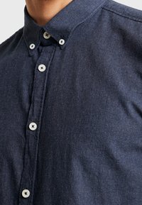 Tummansininen nappipaita, jossa on tekstuuri kangas, klassinen kaulus ja valkoiset napit. Siististi viimeistelty ja tyylikkäästi ommeltu.