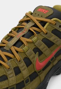 Olīvzaļa un melna Nike sporta apavi ar sarkanu swoosh, oranžiem un dzelteniem svītrotiem šņorēm un "Bowerman Series" tekstu uz mēles cilpas.