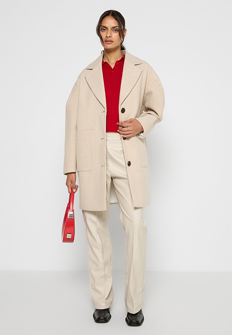 Kobieta w beżowym płaszczu, czerwonym topie, kremowych spodniach, czarnych butach, trzymająca małą czerwoną torebkę, na tle jednolitego białego tła.