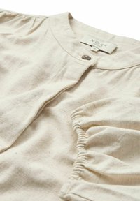 Blusa di cotone beige chiaro con scollatura rotonda, chiusura a singolo bottone, maniche arricciate e una finitura morbida e strutturata.