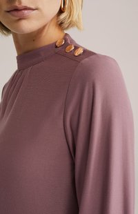 Dugi rukav majica u prigušenoj mauve boji, s visokim izrezom i tri metalna gumba kao ukras na ramenu. Glatka tekstura.