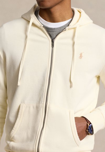 Sweatshirt amarelo com fecho de correr, bolso canguru e cordões, com um pequeno logótipo no peito e textura suave.