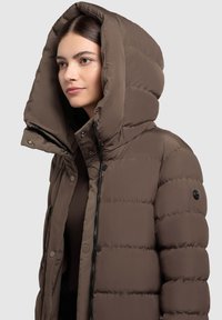 Pruun täispuhutav jope suure ja struktureeritud kapuutsiga, millel on tõmblukk ja trukk kinnitused, sile pind ja keha ümber istuv lõige soonikutega detailidega.