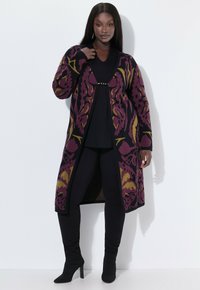 Langer Cardigan in Lila mit floralem Muster in Grün und Schwarz. Aus strukturiertem Strick gefertigt, mit einem Verschluss vorne. Schwarzes Oberteil und Hose darunter getragen.