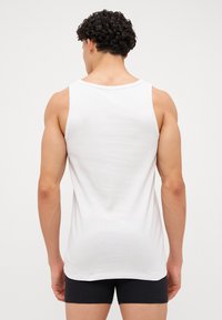 Λευκό αμάνικο μπλουζάκι με στρογγυλή λαιμόκοψη και ραμμένο υλικό, που δείχνει μια λεία πλάτη και ελάχιστες ραφές, σε συνδυασμό με μαύρες βερμούδες.