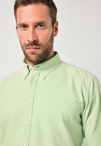 Erwachsener Mann mit kurzen braunen Haaren und Bart, der ein hellgrünes Hemd mit "JP 1880"-Stickerei auf der Brust trägt.