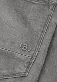 Grå denimtyg med en subtil struktur och en broderad etikett som läser "I.B.J." nära sömmen på bakfickan.