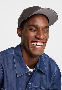 Berretto grigio con visiera curva, indossato da una persona che sorride. Giacca di jeans con colletto e bottoni visibili. Sfondo semplice.