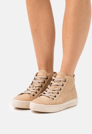 Een paar beige high-top sneakers met crèmekleurige veters gedragen op blote benen, afgebeeld tegen een effen witte achtergrond.