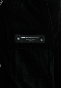 Chaqueta de pana negra con una superficie texturizada, gran bolsillo en el pecho y una etiqueta de cuero con la marca y broches de plata.