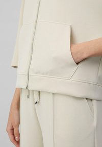 Helles beigefarbenes Zip-Jackett mit einer Fronttasche, aus glattem Stoff gefertigt, mit geripptem Saum und Kordelzugdetails an der Taille.