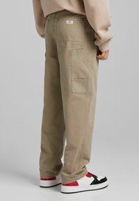 Beige puuvillaiset cargo-housut, joissa on suuret taskut, suora malli ja näkyvät saumat. Yhdistetty punavalkoisiin lenkkareihin.