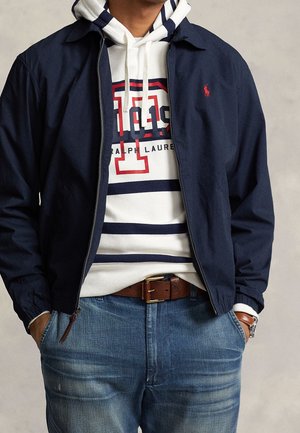 Mann trägt eine marineblaue Jacke, die über einem weißen Hoodie mit rotem und marineblauem Logo offen ist, blaue Jeans, braunen Gürtel und Uhr mit braunem Armband.