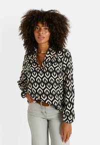 Blusa de patrón negro y crema con un corte suelto, cuello y mangas largas. Pantalones gris claro y un cinturón marrón completan el conjunto.