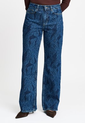 Blauwe wijd uitlopende jeans met grote donkere bloemmotieven, gedragen met bruine puntige schoenen en een bruin topje.