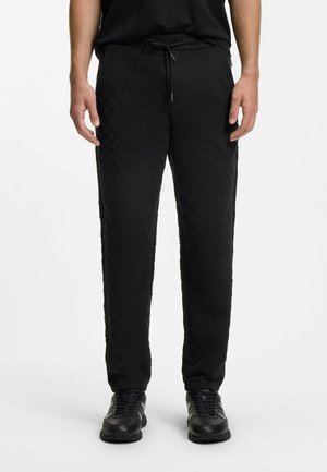 Mann trägt schwarze, strukturierte Jogginghosen mit Kordelzug in der Taille und schwarze Sneakers, steht vor weißem Hintergrund.