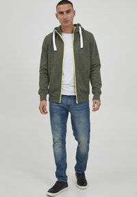 Groene ritssluiting hoodie met witte trekkoorden, voorzien van een gele rits. Gepaard met blauwe spijkerbroek en zwarte sneakers. Gladde textuur, casual ontwerp.
