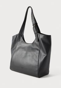 Bolsa tote de couro preta com alça ajustável larga e costura decorativa mínima na superfície lisa, apoiada contra um fundo simples.