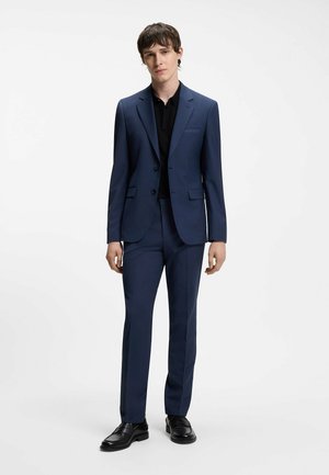 Jeune homme debout portant un costume bleu marine et une chemise noire avec des chaussures habillées noires, sur un fond blanc uni.