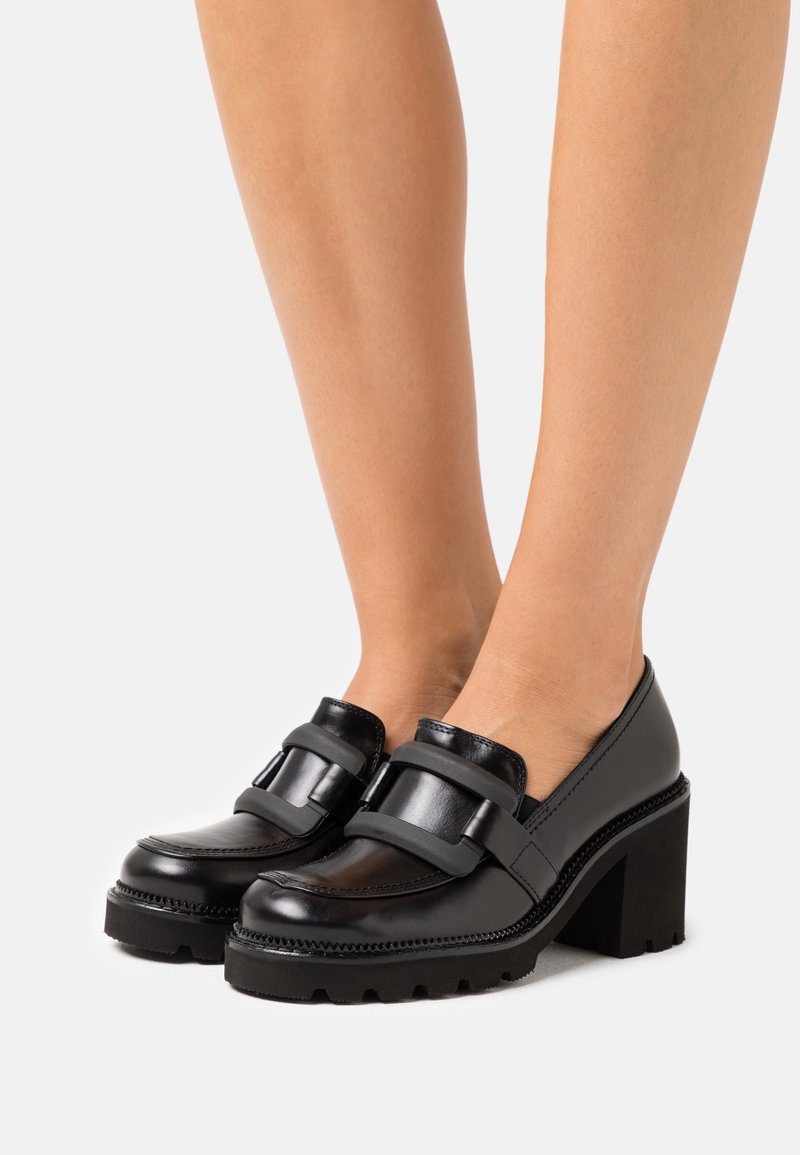 Mocassins en cuir noir avec un talon épais, dotés d'une large bride comme accent. Semelle texturée et coutures détaillées rehaussent le design.