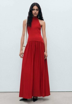 Vestido largo rojo con cuello halter, con un torso ajustado y falda fluida. Textura suave, sin patrones, combinado con zapatos negros de punta y un bolso crema.