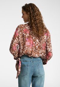 Donna con capelli ricci che indossa un blusa a fiori marrone legata in vita e jeans blu a vita alta, mostrata di spalle.